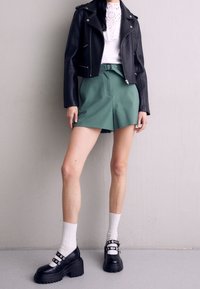 Giacca da biker in pelle nera, top in pizzo bianco, pantaloni corti verdi a vita alta e scarpe con zeppa nere con dettagli argentati. Calzini bianchi alla caviglia visibili.