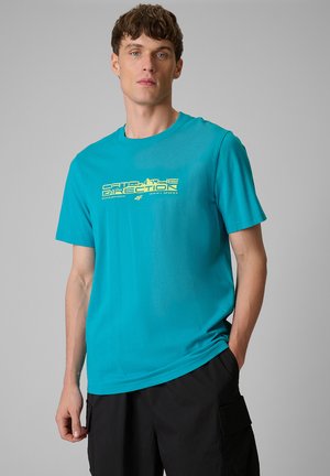 Młody mężczyzna ubrany w turkusowy t-shirt z żółtym napisem "Catch the Direction" i czarne szorty cargo, stoi na jednolitym szarym tle.