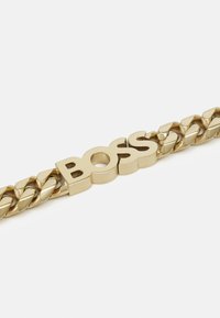 Pulsera de cadena de oro con un diseño retorcido y un logo "BOSS" en el centro. El material tiene un acabado brillante y suave.
