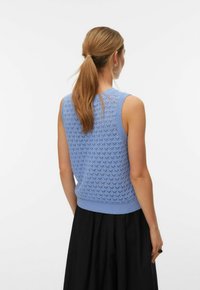 Femme avec une queue de cheval châtain clair portant un haut tricoté sans manches bleu clair et une jupe noire, vue de dos sur un fond uni.