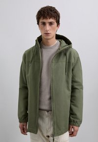 Un jeune homme portant une veste à capuche vert olive par-dessus un pull beige et un pantalon crème, se tenant devant un fond clair.