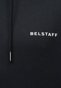 Μαύρο ύφασμα με λευκό κείμενο "BELSTAFF" και μαύρο κορδόνι με ασημένια μεταλλική άκρη ορατή στην αριστερή πλευρά.