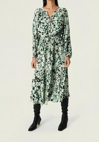Vestido de estampado floral en verde, negro y crema, con mangas largas, cintura ajustable con cordón y diseño de falda en capas. Textura de tela suave.