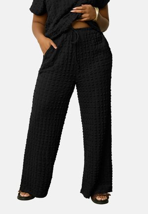 Frau trägt strukturierte schwarze weite Hosen mit Kordelzug in der Taille und schwarze Sandalen, eine Hand in der Tasche, kurz geschnittenes Oberteil leicht angehoben.