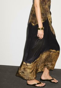 Robe maxi en tissu noir lisse, avec un motif dégradé doré sur l'ourlet. Associée à des tongs marron et un bracelet doré.