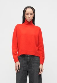Czerwony sweter z golfem o luźnym kroju, dzianinowej fakturze i prążkowanym wykończeniu na dole. W zestawie z wysokim stanem ciemnoszarymi dżinsami.