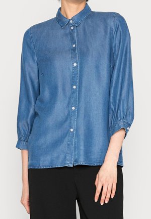 Femme portant une chemise en jean bleu à manches longues boutonnée avec col et un pantalon noir sur un fond uni.