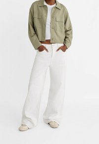 Przycięta oliwkowozielona kurtka z guzikami z przodu, dwoma kieszeniami na piersi; w połączeniu z szerokimi białymi dżinsami i beżowymi sneakersami.