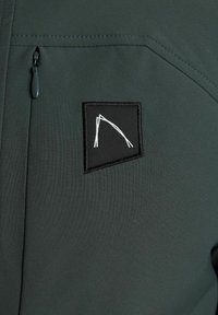 Chaqueta de concha blanda verde oscuro con un bolsillo con cremallera y un parche negro con diseño geométrico blanco. Tejido suave y texturizado.