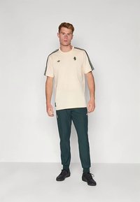 Hele beež värvi T-särk mustade triipude ja embleemiga õlgadel, sobitatud tumerohelistest pükstest. Mõlemal rõivaesemel on lõdvestunud lõige ja elastsed detailid.