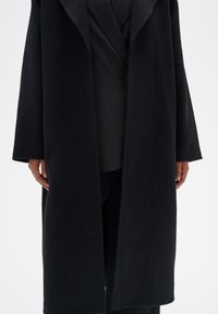 Manteau en laine noir avec un large col, des manches longues et une coupe droite. La texture semble douce, avec un design minimaliste et sans fermetures visibles.