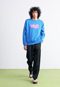 Blå sweatshirt med pink "Nike" logo, parret med sorte sweatpants. Modellen bærer sorte og pink sportsko på et grønt gulv.