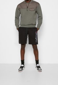 Uomo con felpa oliva e marrone, pantaloni corti Champion neri, calze nere e sneakers bianco e nere, in piedi con le mani in tasca.