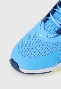 Blå sportssko med ett mesh-övre, texturerade Accenter och en vit sula med en gul Nike-logotyp på sidan. Snörningen är blå.