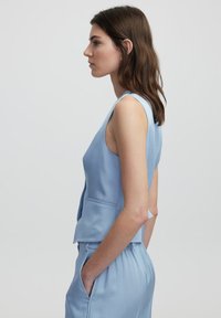 Top senza maniche di un blu chiaro con scollatura a V e design aderente; presenta tasche laterali e una texture liscia. Abbinato a pantaloni coordinati.