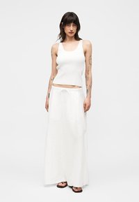 Femme avec des tatouages portant un haut blanc sans manches, une longue jupe blanche avec taille à cordon et des sandales noires, debout devant un fond uni.