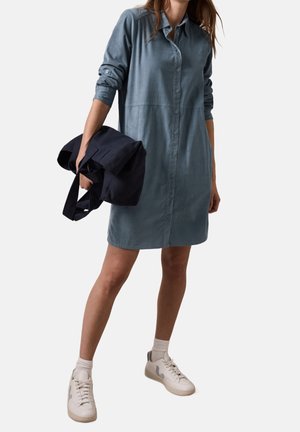 Kobieta ubrana w szarą sukienkę koszulową na długi rękaw, trzyma złożoną czarną kurtkę i ma na sobie białe trampki z białymi skarpetkami.