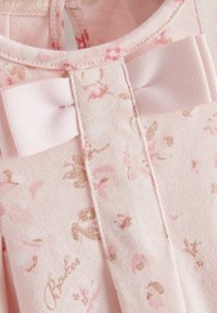 Lähikuva vaaleanpunaisesta kankaasta, jossa on kimaltava kukkakuosi ja teksturoitu vaaleanpunainen nauhakirjaus kaula-aukon lähellä.