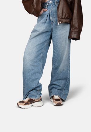 Personne portant un jean large bleu clair, des baskets marron et beige, et une veste courte en cuir marron avec la main dans la poche.