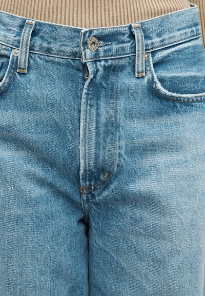 Κοντινό πλάνο στη μέση ενός ανοιχτόχρωμου τζιν με μεταλλικό κουμπί, θηλιές ζώνης και μερικώς ανοιχτό φερμουάρ, φορεμένο με μπεζ ριμπ πουλοβεράκι.