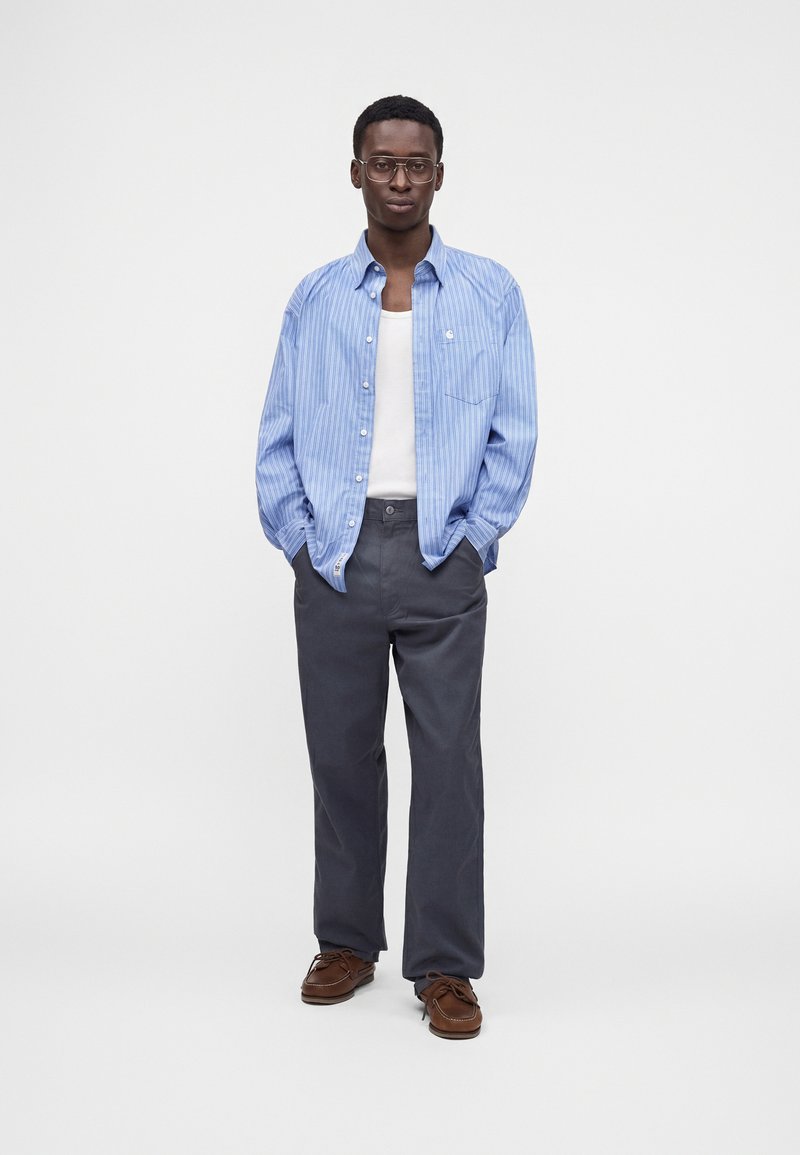 Homme debout les mains dans les poches portant des lunettes, une chemise bleue rayée sur un débardeur blanc, un pantalon foncé et des chaussures marron.