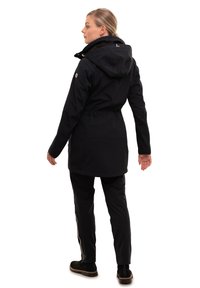 Veste noire imperméable avec capuche, taille élastique et poches zippées. Associée à un pantalon noir avec des bandes blanches sur les côtés et des chaussures robustes.