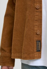 Braune Cordjacke mit Knopfverschlüssen, geriffelter Textur und einem Markenlabel am Saum. Getragen über einem weißen Shirt.