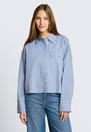 Młoda kobieta ubrana w luźną, niebieską koszulę w paski z dużymi mankietami oraz niebieskie dżinsy, stojąca na jednolitym tle.