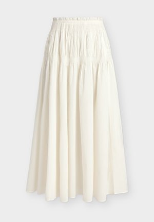 Femme aux cheveux courts naturels portant une chemise crème boutonnée nouée, une longue jupe crème, des sandales beiges et tenant un petit sac à main marron.