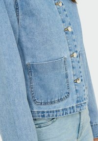 Veste en denim bleu clair au design ajusté, avec des boutons en métal argenté et deux poches avant. Possède des détails de couture et une longueur légèrement raccourcie.