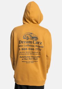 Gele hoodie met een grote graphic featuring een illustratie van een vintage auto en tekst voor "Dream Carz" autoservice op de achterkant.