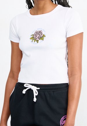 Femme portant un t-shirt blanc ajusté avec un motif de rose rose et du texte, associé à un pantalon noir à cordon avec un logo circulaire rose.
