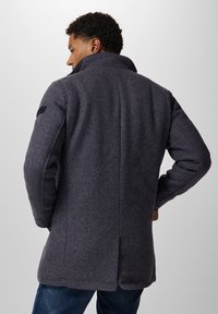 Cappotto in misto lana grigio scuro con colletto alto, vestibilità sagomata e uno spacco posteriore. Presenta un tessuto testurizzato e una toppa sulle spalle.