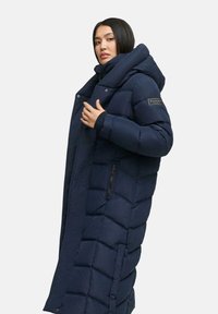 Geacă puffy de un albastru marin, cu un design lung și structurat, glugă supradimensionată, buzunare cu fermoar și un patch cu logo pe brațul stâng.