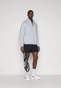 Felpa grigio chiaro con zip e maniche lunghe, abbinata a pantaloncini neri. Il modello tiene in mano una racchetta da tennis e indossa sneakers bianche.