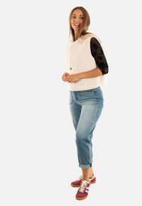 Fleece vest in licht crème met een ronde kraag en knoopsluiting; gecombineerd met lichtblauwe opgerolde jeans en bordeauxrode sneakers met witte accenten.