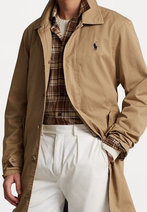 Uomo che indossa un trench beige sopra una camicia a quadri marrone e pantaloni bianchi plissettati, mano nella tasca del cappotto, volto non visibile.