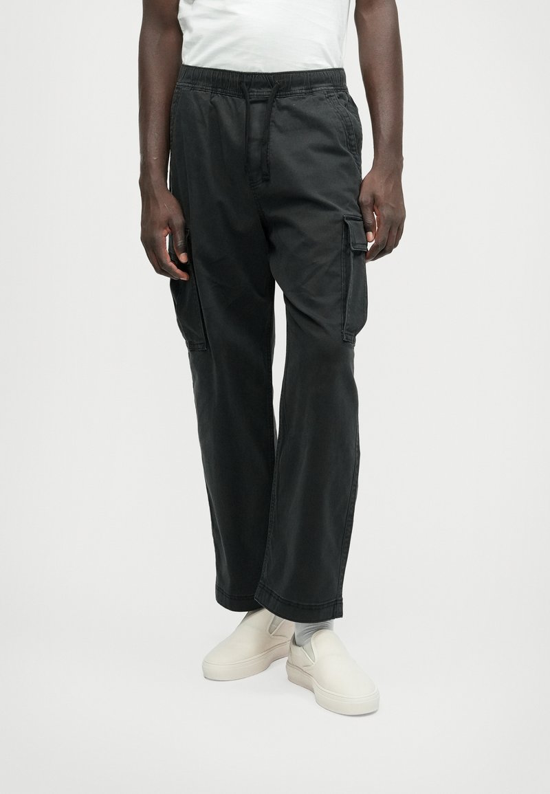 Melni kokvilnas kravas bikses ar elastīgu jostu, sānu kabatām un brīvu piegriezumu, kopā ar baltām slip-on kurpēm.