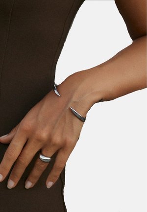 Silberner Metallring und Armband mit einem eleganten, geschwungenen Design und spitzen Akzenten. Glatte Textur auf einem gebräunten Hautton.