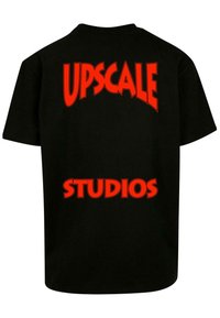 Čierne bavlnené tričko s výrazným červeným textom na chrbte: "UPSCALE" klenuté nad "STUDIOS," s klasickým dizajnom okrúhleho výstrihu.