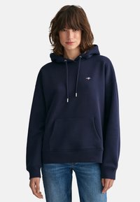 Granatowa bluza z kapturem z paskami do ściągania, przednią kieszenią i ściągaczami na mankietach. Posiada niewielkie logo na klatce piersiowej. Miękka, teksturowana tkanina.
