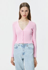 Cardigan a maniche lunghe ribbed rosa chiaro con scollatura a V e chiusura a bottoni, abbinato a jeans blu chiaro a vita alta.