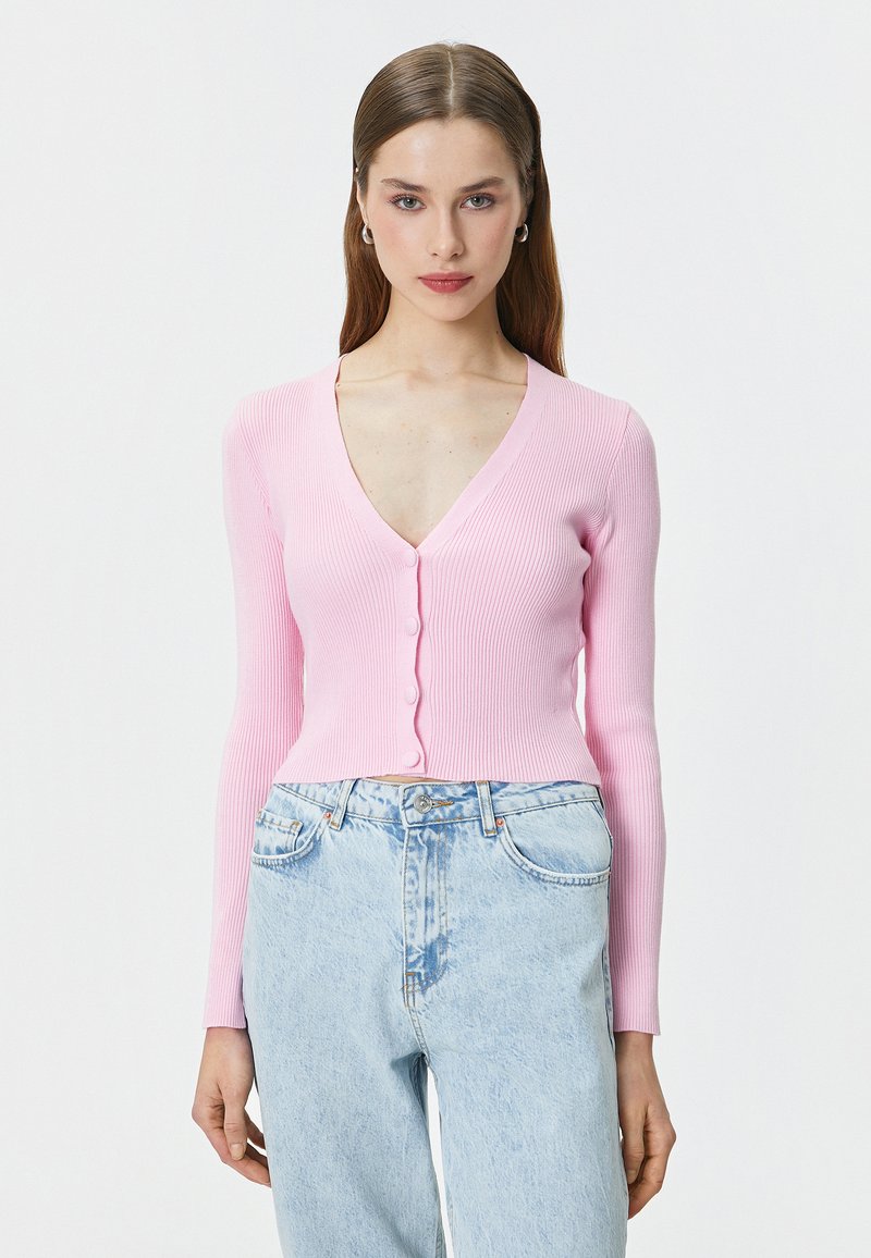 Cardigan a maniche lunghe ribbed rosa chiaro con scollatura a V e chiusura a bottoni, abbinato a jeans blu chiaro a vita alta.