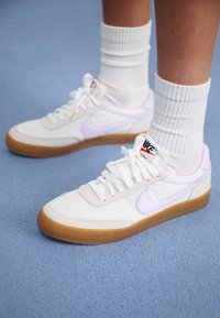 Adidași albi din pânză cu un swoosh Nike violet deschis, talpă din cauciuc și șireturi plate. Purtați cu ciorapi albi cu dungi, pe un fundal albastru.