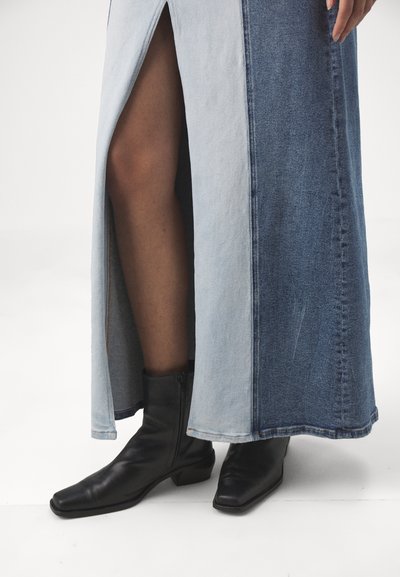 Denim maxi skirt featuring a high slit, with contrasting light and dark blue sections, paired with black leather ankle boots.