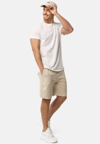 Let sval, kortærmet hvid T-shirt med et lille logo, parret med beige shorts og hvide sneakers. Model iført en beige kasket.