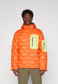 Dikke oranje jas met een gestructureerde buitenkant, teal ritsen en een groene ritszak op de linkerborst. Voorzien van een capuchon en elastische manchetten.