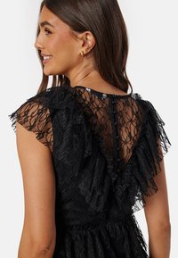 Robe en dentelle noire avec des manches courtes à volants et un dos transparent. Présente des boutons le long du dos central et une taille ajustée.