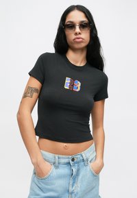 Γυναίκα με μακριά σκούρα μαλλιά και γυαλιά ηλίου, φορώντας ένα μαύρο κοντό t-shirt με πολύχρωμη εκτύπωση "23" και ανοιχτό μπλε τζιν, με τα χέρια στις τσέπες.