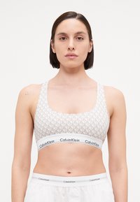 Svijetlosivi sportski grudnjak s umirujućim uzorkom logotipa, s širokim bijelim pojasom na kojem je ime "Calvin Klein" i podesivim naramenicama.